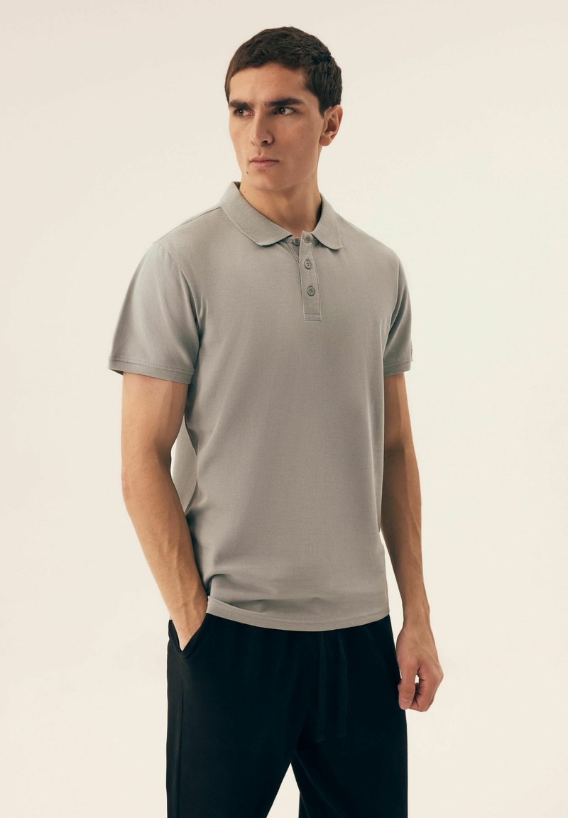 Szara koszulka polo z klasycznym kołnierzykiem, zapięciem na trzy guziki i krótkimi rękawami, zestawiona z czarnymi spodniami. Gładka tekstura materiału.