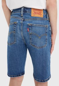 Modré denimové šortky s klasickým střihem, pěti kapsami a koženou nášivkou na pase s detaily značky. Viditelné kontrastní švy.
