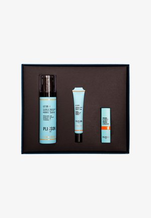 Three skincare products in light blue packaging are displayed in a brown box. The largest bottle is cylindrical, the others are tapered tubes.