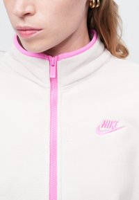 Nike dukserica od flisa u svijetloj krem boji s ružičastim zatvaračem i naglascima na ovratniku; sadrži mali ružičasti logo na prsima. Mekana tekstura, ležeran dizajn.