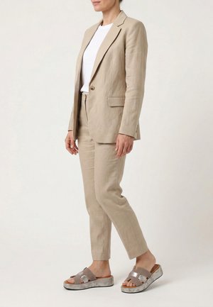 Person iført beige skræddersyet buksedragt med enkeltknappet blazer, hvid skjorte og taupe platforms-sandaler mod ensfarvet baggrund.