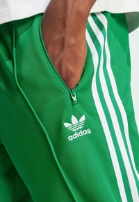 Pantalones de chándal verdes de poliéster con diseño de tres rayas blancas a lo largo del costado, detalles de bolsillo con cremallera y un logo de Adidas bordado.