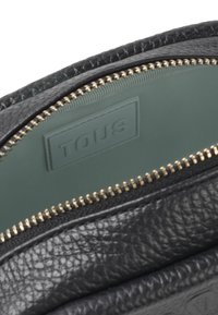 Czarna skórzana torba z zamkiem błyskawicznym, z zielonym wykończeniem wnętrza i teksturowanym zewnętrzem. Zawiera widoczną, wytłoczoną etykietę "TOUS".