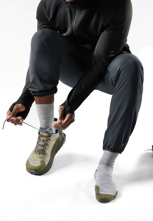 Person crouching while tying shoelaces on beige and olive sneaker, wearing gray socks and dark athletic pants and jacket.