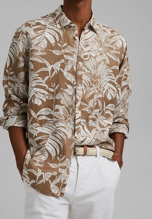 Hombre con camisa marrón abotonada con estampado blanco de hojas tropicales, pantalones blancos, cinturón blanco trenzado con detalles de cuero marrón, manos en los bolsillos.