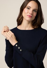 Granatowy sweter wykonany z miękkiego materiału, z okrągłym dekoltem i guzikami na rękawach. Gładka faktura z prążkowanymi mankietami.