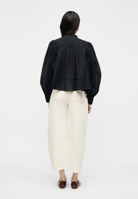 Γυναίκα με μακριά σκούρα μαλλιά, φορώντας φαρδιά μαύρη μπλούζα, εκρού παντελόνι με φαρδιά μπατζάκια και σκούρα καφέ παπούτσια, στέκεται μπροστά σε λευκό φόντο.