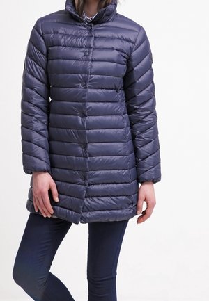 Femme portant une doudoune matelassée bleu marine avec des boutons-pression, associée à un pantalon foncé, debout devant un fond uni.