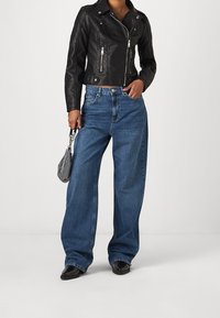 Czarna skórzana krótka kurtka motocyklowa, niebieskie dżinsy z wysokim stanem i szerokimi nogawkami, oraz mała szara torebka o teksturowanej powierzchni z łańcuszkowym paskiem. Czarne obuwie.