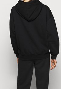 Haut en molleton noir surdimensionné avec capuche à cordon de serrage, épaules tombantes, poignets côtelés et tissu texturé. Associé à un pantalon noir assorti.