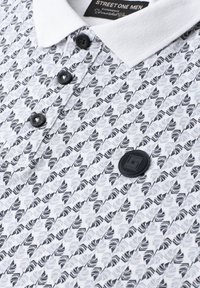 Polo blanc avec motif géométrique noir, boutons noirs, col blanc et un patch logo rond noir sur la poitrine.