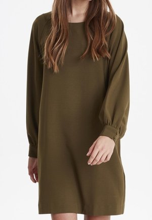 Woman wearing a loose olive green long-sleeve dress with gathered cuffs, standing against a plain white background.