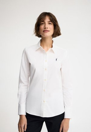 Frau mit kurzen braunen Haaren, die ein weißes, zum Knöpfen geöffnetes Hemd mit einer kleinen dunklen Stickerei auf der linken Brust trägt, kombiniert mit schwarzen Hosen.