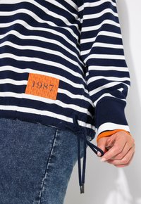 Sweatshirt rayé bleu marine et blanc avec un cordon de serrage à l'ourlet. Présente une étiquette orange avec "1987" et des poignets côtelés en orange et blanc.