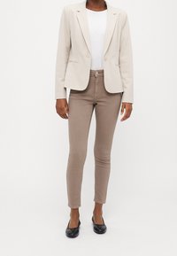 Mujer vestida con un blazer beige, camisa blanca, pantalones ajustados topo y bailarinas negras, de pie contra un fondo claro y liso.