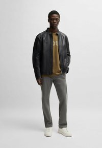 Giubbotto bomber in pelle nera con colletto a coste, t-shirt verde oliva con testo del logo, pantaloni grigi e sneakers bianche.