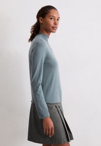 Lichtblauw langeklauwenshirt met een hoge hals, gecombineerd met een grijze geplooide rok. De stof lijkt zacht en glad.