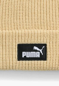 Pletená béžová čiapka s ryhovanou textúrou; obsahuje čiernu obdĺžnikovú nášivku s bielym označením "PUMA" a skáčúcim logom pumu.