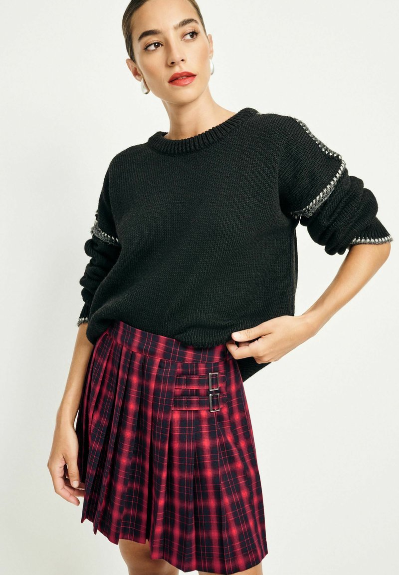 Black knit sweater with rolled sleeves, paired with a red and black plaid skirt featuring pleats and two buckle accents.