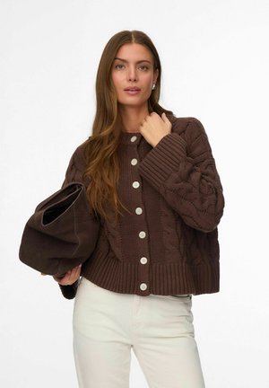 Femme portant un cardigan marron à torsades boutonné, tenant un grand sac marron foncé, et portant un pantalon blanc sur un fond uni.