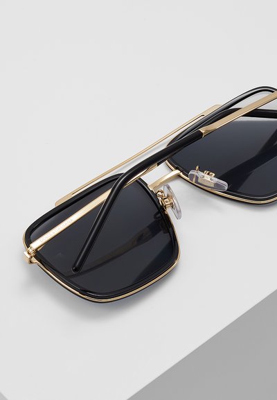 Gafas de sol rectangulares con lentes negras, montura fina dorada y varillas negras descansando sobre una superficie blanca.