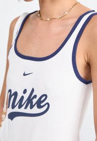 Vit ribbad linne med marinfärgad kant, med en stor marinfärgad "Nike"-logotyp på framsidan. Slät textur, casual design.