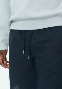 Gros plan d'une personne portant un pull léger et un short bleu marine foncé avec cordon de serrage, montrant la main gauche détendue sur le côté.