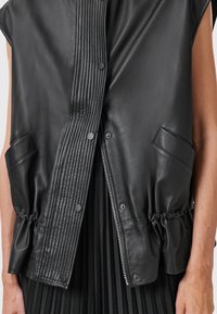 Black leather vest featuring a smooth finish, ribbed front panel, snap closures, elasticized waist, and two side pockets.