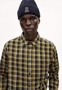 Geruite shirt in geel en marineblauw, gemaakt van een stof met een gladde textuur; heeft een button-down kraag en een borstzak. Gebreide marineblauwe beanie op het hoofd.