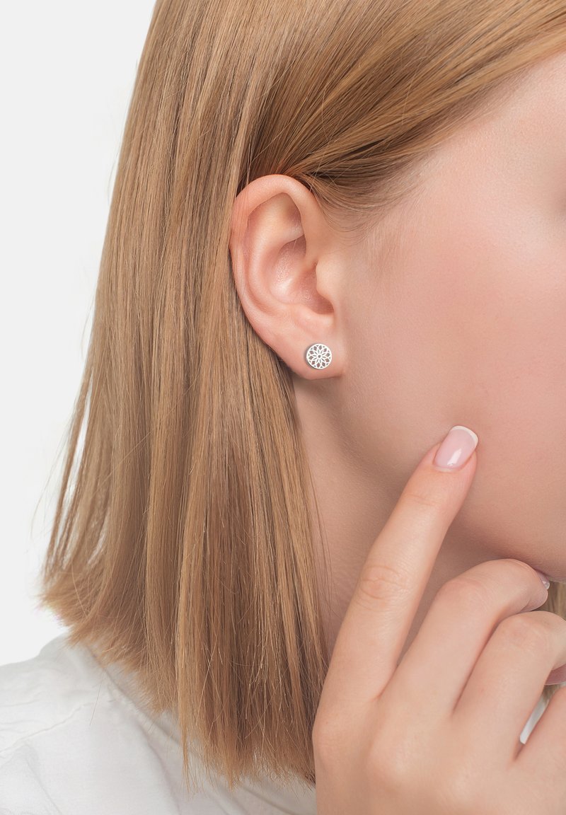 Runda silverfärgade stiftörhängen med ett utskuret blommönster, bärs i örat, tillsammans med rakt blont hår och en ljusfärgad skjorta.