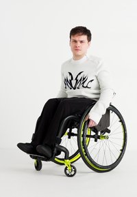 Black and white patterned sweatshirt, black pants, seated in a wheelchair with vibrant yellow accents and black wheels, against a white background.