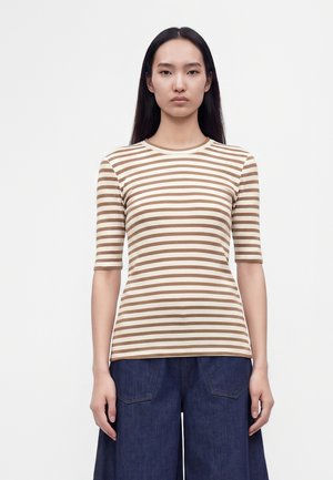 Femme aux longs cheveux noirs portant une chemise beige et blanche à rayures avec des manches aux coudes et un pantalon large bleu foncé.