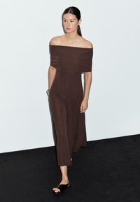 Robe brune à épaules dénudées au design ajusté, avec un décolleté droit et une longueur mi-mollet. Texture lisse, portée avec des sandales noires à enfiler.