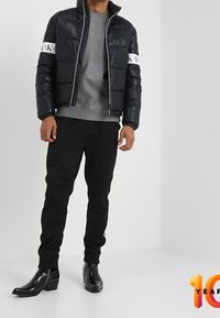 Giacca puffer nera con colletto alto e strisce logo bianche, abbinata a una felpa grigia e pantaloni neri, completata da stivali neri lucidi.
