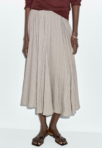 En beige, plisserad midikjol gjord av texturerat tyg, med vertikala ränder och en flödande silhuett. Matchad med bruna sandaler.