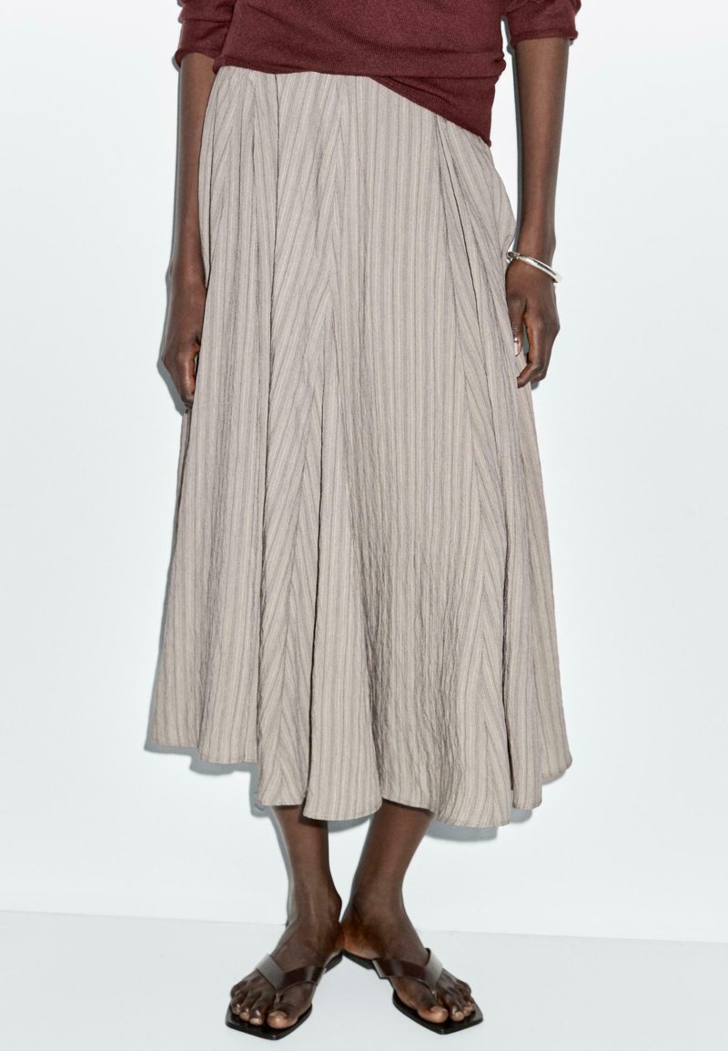 Een beige, geplooide midi-rok gemaakt van textiel met een gestructureerde stof, voorzien van verticale strepen en een vloeiende silhouet. Gecombineerd met bruine sandalen.