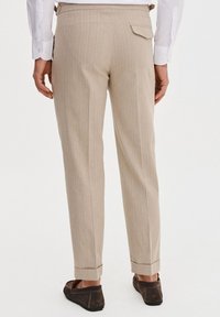 Beżowe spodnie w paski, wykonane z lekkiego materiału, z pojedynczą tylną kieszenią i podwiniętymi nogawkami. Połączone z ciemnymi butami.
