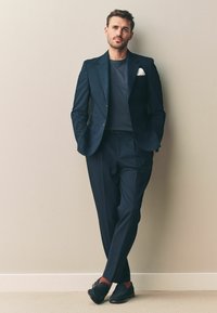 Marineblå jakkesæt med en tætsiddende blazer og plisserede bukser, parret med en grå skjorte og sorte loafers, akkompagneret af et hvidt lommetørklæde.