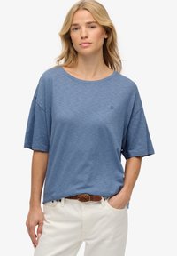 Lichtblauw kortekoope shirt, losse pasvorm met een ronde hals, met een subtiele textuur en een klein logo op de borst. Gepaard met crèmekleurige broeken.