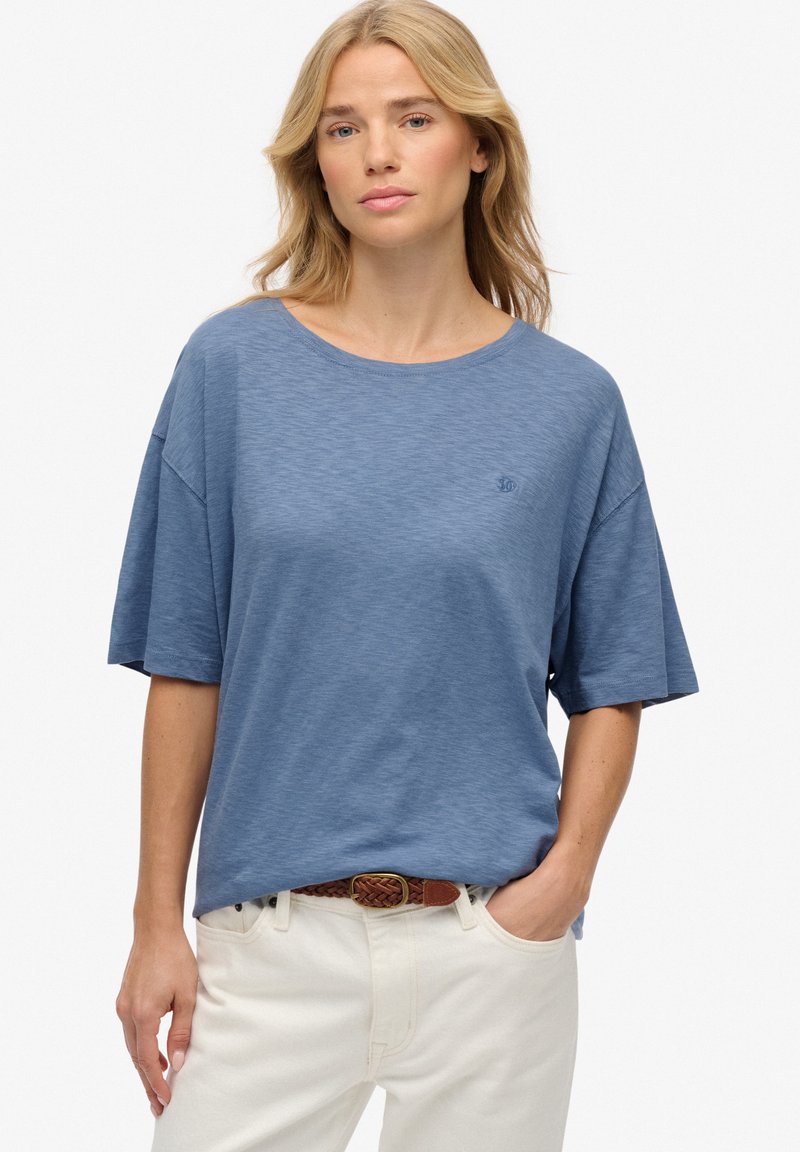 Lichtblauw kortekoope shirt, losse pasvorm met een ronde hals, met een subtiele textuur en een klein logo op de borst. Gepaard met crèmekleurige broeken.