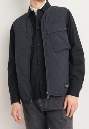 Mężczyzna noszący czarną pikowaną kamizelkę na suwak, czarną koszulę zapinaną na guziki i ciemnoszare dżinsy, stojący na jednolitym tle.