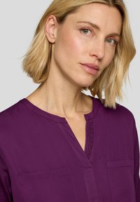 Lila Bluse mit V-Ausschnitt und dekorativem Beading entlang der Mitte, zwei Fronttaschen und einer glatten Stofftextur.