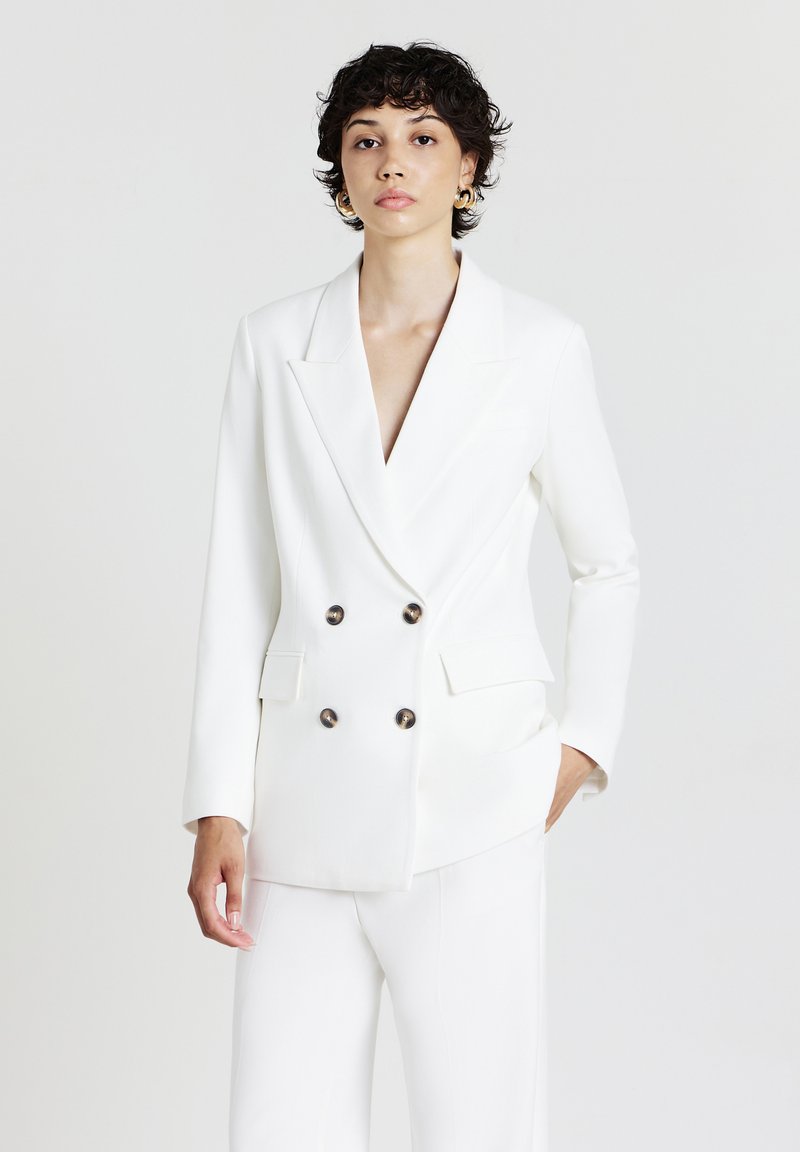 Blazer bianco a doppio petto con rever affilato, caratterizzato da bottoni rotondi neri e vestibilità strutturata. Tessuto liscio con design sartoriale.