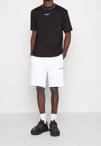 Maglietta nera con logo bianco, pantaloncini bianchi con dettaglio logo e sneakers nere con lacci bianchi. Outfit casual con vestibilità comoda.