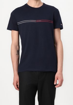 Mężczyzna ubrany w granatową koszulkę Tommy Jeans z czerwono-białymi poziomymi paskami i czarne spodnie, stojący na białym tle.