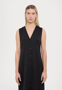Rochie neagră fără mâneci, cu un decolteu adânc în V, având închidere frontală cu nasturi și un material neted și lucios. Design simplu și elegant.