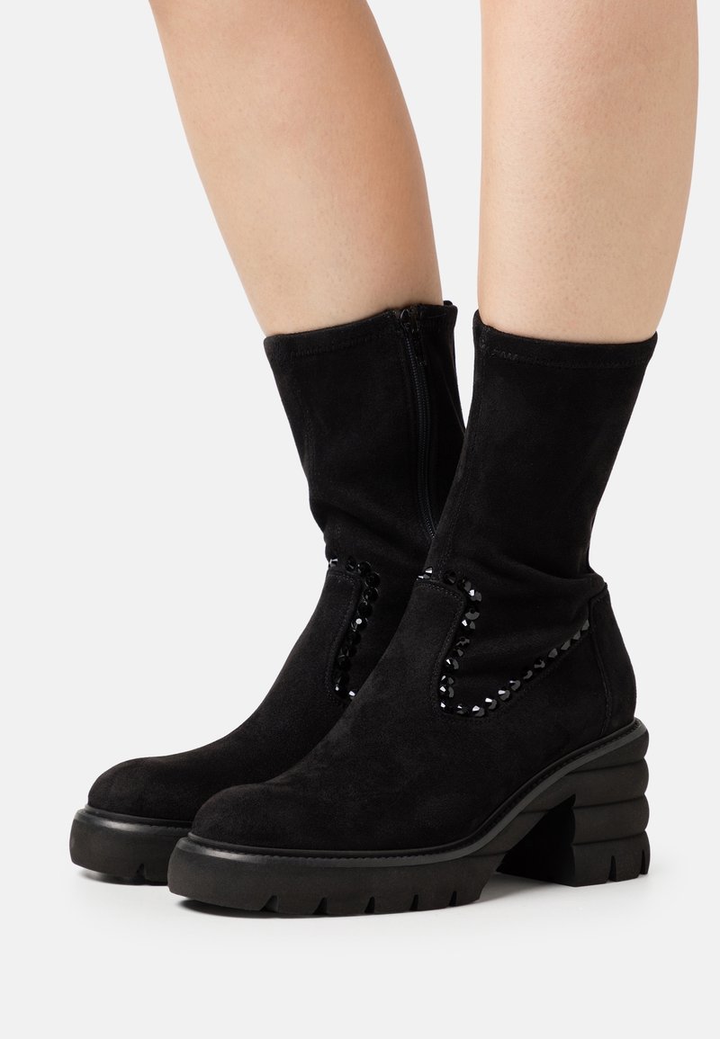 Bottes en daim noir montant à la cheville avec talons épais et carrés, fermetures éclair latérales, et ornements en sequins noirs, portées sur des jambes nues.