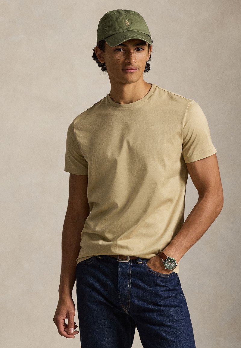 Μπεζ κοντομάνικη μπλούζα, μαλακό β αμβακερό υλικό, φορεμένη με σκούρο τζιν. Πράσινο καπέλο, ανθεκτικό στο νερό ρολόι με καφέ λουράκι, casual εφαρμογή.