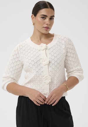 Žena oblečená v bielom pletenom kardigáne s kvetinovými gombíkmi, čiernymi nohavicami, zlatými kruhovými náušnicami a zlatým náramkom pózuje so zloženými rukami.