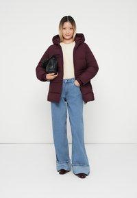 Burgundy puffer jacket with a hood, cream sweater, wide-leg blue jeans, and a black drawstring bag; plain background.
